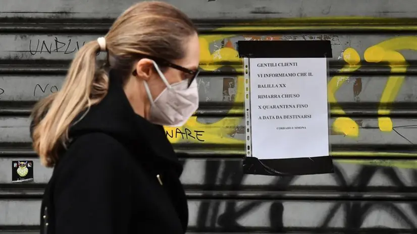 Una passante con mascherina davanti alla saracinesca abbassata di un bar chiuso per quarantena - Foto Ansa © www.giornaledibrescia.it