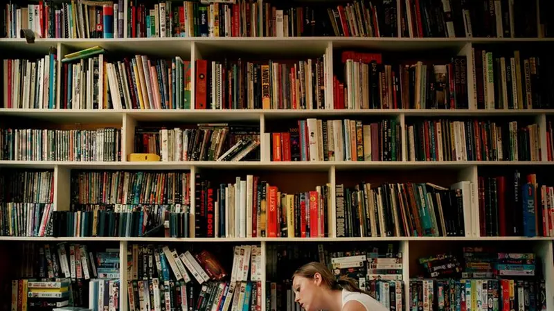 Young woman lying on floor by book shelves, reading, side view
