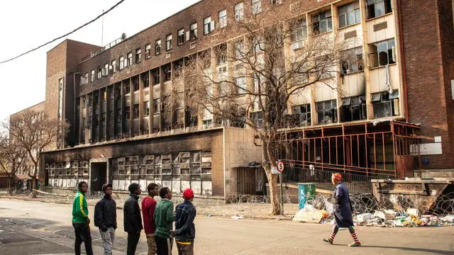 epa10832893 People stand in front of the gutted 80 Albert Street building, where at least 73 people died in a fire, in downtown Johannesburg, South Africa, 01 September 2023. According to Emergency Management Services spokesman Robert Mulaudzi, at least 73 people have died and 52 people were injured in a building fire early morning on 31 August. An investigation into the cause of the fire has been opened as mopping-up operations continue throughout the day. EPA/KIM LUDBROOK