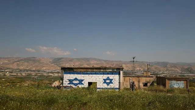 epa11214811 An Israeli right-wing activist walks past an abandoned West Bank house after painting it with an Israeli national flag, in the Jordan Valley, as the city of Ajlun, Jordan, is seen in the background, 11 March 2024. EPA/ATEF SAFADI