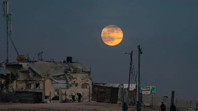 epa11779006 Displaced people walk near destroyed buildings as the full moon rises over Khan Yunis, southern Gaza Strip, 15 December 2024. According to the UN, at least 1.9 million people (or nine in ten people) across the Gaza Strip are internally displaced, including people who have been repeatedly displaced. EPA/HAITHAM IMAD