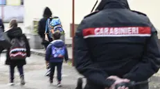 Bambini con gli zaini vanno verso la scuola media di Scanzano mentre un carabiniere li segue con lo sguardo, a Castellammare di Stabia, 18 novembre 2024. I militari presidiano l'istituto per prevenire tensioni in occasione della ripresa delle lezioni, dopo il caos suscitato dall'aggressione di una trentina di genitori ai danni di una insegnante di sostegno. La docente aggredita, a casa dopo il trauma cranico riportato, non è ovviamente in servizio ma le famiglie che la contestano, dando credito alle voci su presunte molestie sessuali ai danni di alcuni alunni, insistono nel chiedere il suo allontanamento e minacciano di far disertare le lezioni ai propri figli. ANSA / CIRO FUSCO (simbolica, generica, carabiniere, carabinieri, cc, scuola, studenti, controlli, violenza, denuncia)