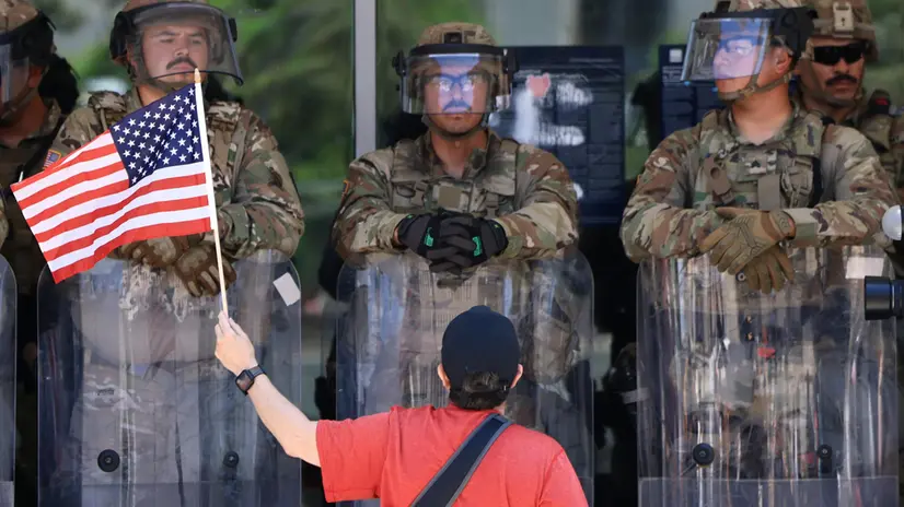 epa12216604 A protester with a US flag stands in front of US Marines guarding the Edward R. Roybal Federal Building, during an Anti-ICE protest held during the Fourth of July holiday, in Los Angeles, California, USA, 04 July 2025. EPA/DAVID SWANSON