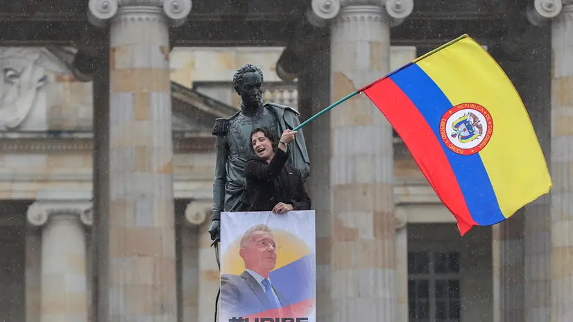 epa12288807 A person holds a flag and a placard during a demonstration in support of former President Alvaro Uribe at the Plaza de Bolivar in Bogota, Colombia, 07 August 2025. Thousands of people began gathering in the streets of Colombia's major cities to show their support for Uribe, who was sentenced to 12 years of house arrest for procedural fraud and bribery in criminal proceedings, and at the same time to reject the government of the current president, Gustavo Petro. EPA/CARLOS ORTEGA