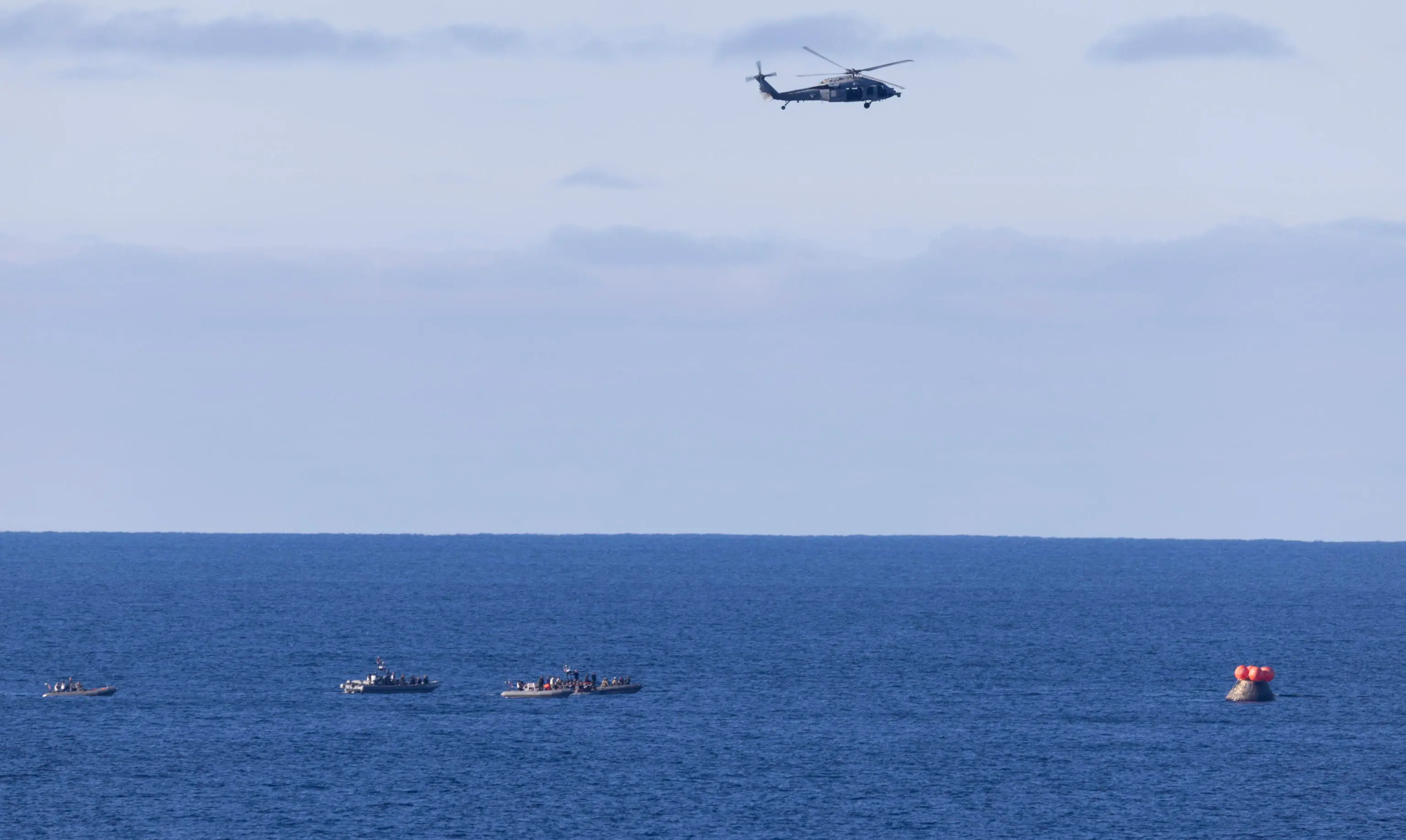 epa12881926 A handout photo made available by the National Aeronautics and Space Administration (NASA) shows a US Navy MH-60 Seahawk from Helicopter Sea Combat Squadron (HSC) 23 flying overhead as small boats approach NASA's Orion spacecraft with Artemis II crewmembers after splashdown in the Pacific Ocean, 10 April 2026. The mission, crewed by NASA astronauts Reid Wiseman, commander, Victor Glover, pilot, Christina Koch, mission specialist, and CSA (Canadian Space Agency) astronaut Jeremy Hansen, mission specialist, completed a flyby of the Moon and returned to Earth. EPA/JOEL KOWSKY / NASA / HANDOUT HANDOUT EDITORIAL USE ONLY/NO SALES